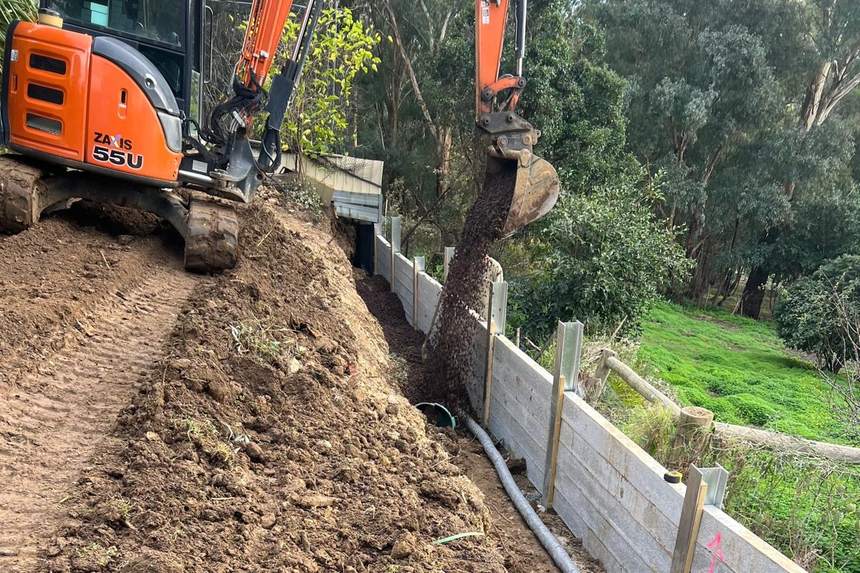 Excavator dropping scoria into the trench behind the retaining wall over the agi drain — Templestowe