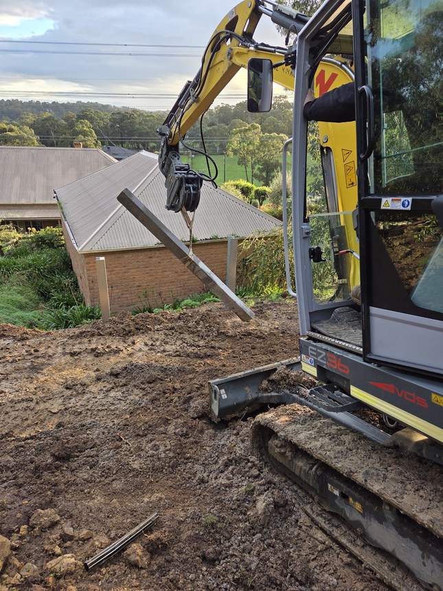 Excavator lifting a 50MPa concrete sleeper into place between the steel posts — Templestowe